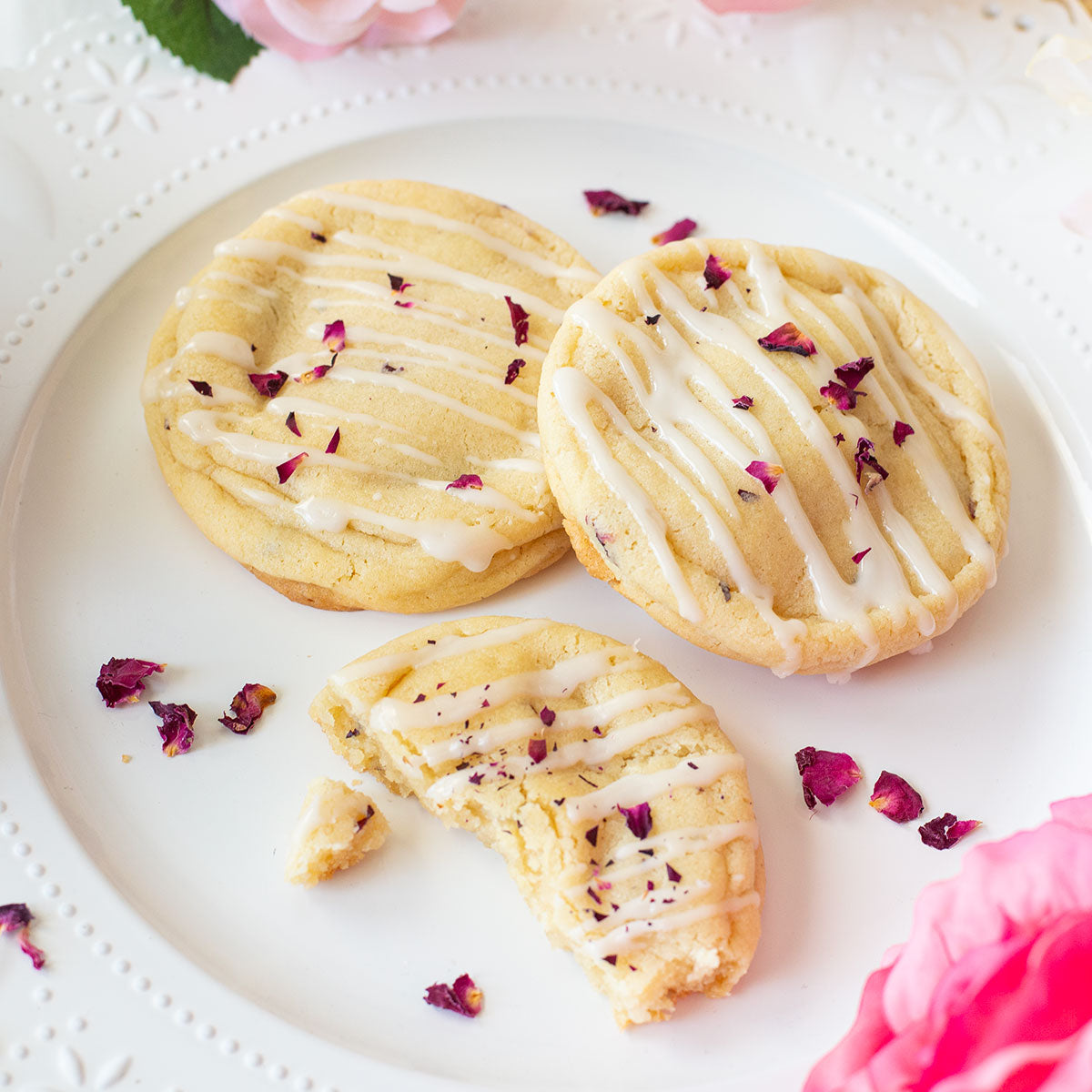 Three Champagne & Roses Cookie sits elegantly on a plate, adorned with fresh rose petals, emphasizing its delicate champagne and rose water flavor