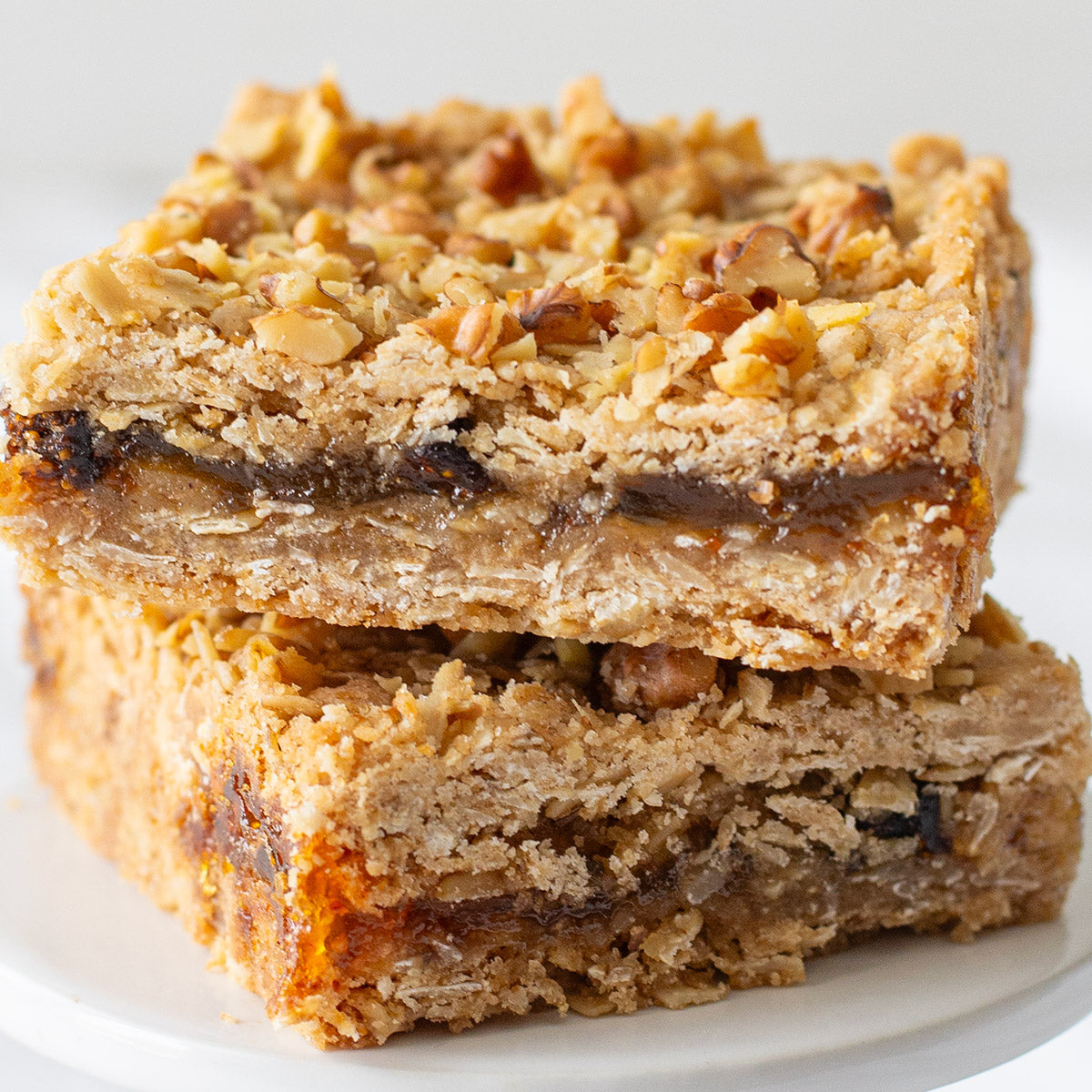 A close-up of two Figgy Walnut Bars stacked on top of each other, revealing the gooey fig layer nestled between a rich walnut crumble topping and a buttery brown sugar and oat crust, highlighting their chewy texture and inviting fall flavors.