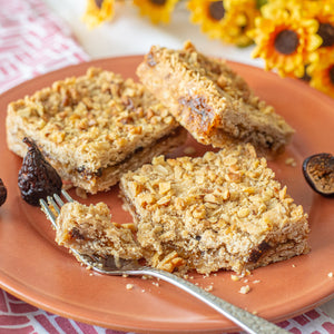 Three Figgy Walnut Bars sit on a plate, one with a piece cut out using a fork, surrounded by dried figs. In the background, fall flowers add a cozy and inviting seasonal touch.