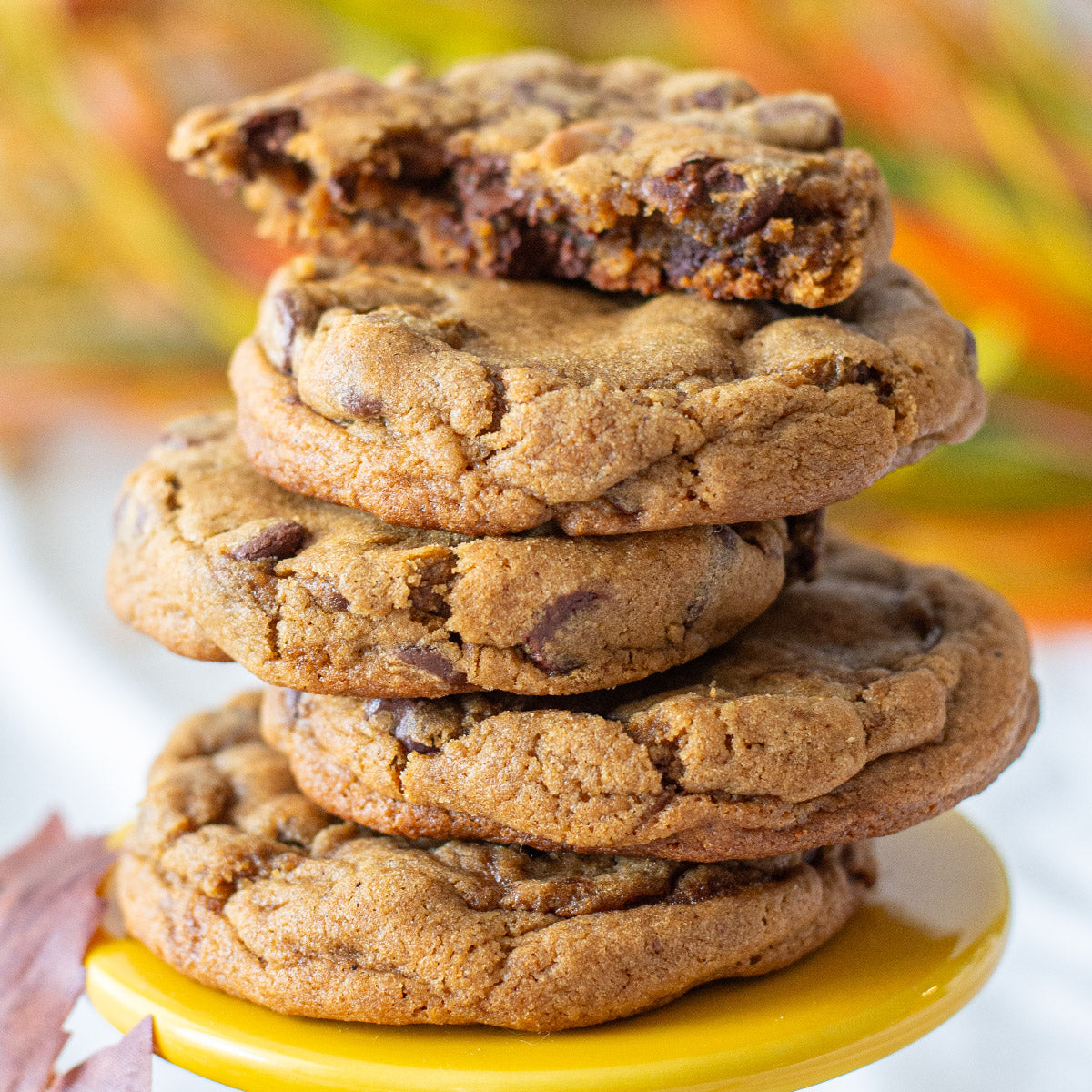 A stack of five Harvest Spice Chocolate Chip Cookies, with the top cookie broken in half to showcase its gooey, rich interior filled with chocolate chips and fragrant spices. The golden-brown cookies exude warmth and coziness, complemented by a vibrant fall leaf and colorful autumn foliage in the background, enhancing the seasonal theme.