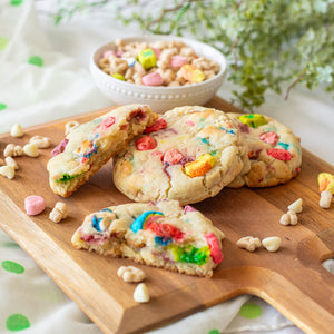 Wooden board with Lots 'O Luck cookies surrounded by cereal and white chocolate chips, and a bowl of cereal in the background.