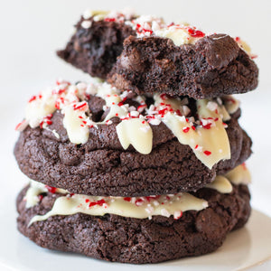 Close-up of a stack of Peppermint Mocha Cookies. The cookies feature rich chocolate dough drizzled with white chocolate and topped with crushed peppermint candy. The top cookie is broken in half, revealing its fudgy interior and chocolate chips. Perfect for holiday indulgence.