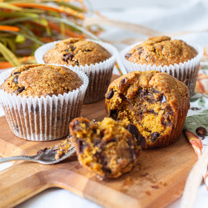 Four Pumpkin Chocolate Chip Muffins are arranged on a surface, with one muffin removed from its cup and broken in half to reveal the moist, spiced pumpkin interior. Visible chunks of semi-sweet and milk chocolate chips are nestled throughout the soft, orange crumb, highlighting the warm, autumnal flavors.