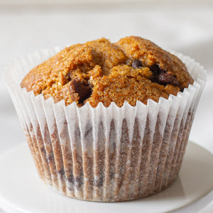 A single Pumpkin Chocolate Chip Muffin sits upright, its top domed and slightly cracked, revealing chunks of semi-sweet and milk chocolate chips. The warm orange hue of the pumpkin contrasts with the speckled chocolate, making it look moist and freshly baked.