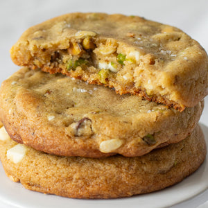 A stack of Salted Honey Pistachio Cookies, highlighting their golden-brown exterior, with visible chunks of pistachios and white chocolate chips peeking through. The cookies are topped with a sprinkling of sea salt flakes, adding a touch of texture and flavor.