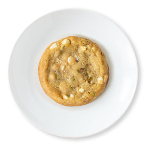 A close-up overhead shot of a Salted Honey Pistachio Cookie, showcasing its golden-brown edges, scattered salt flakes, and chunks of pistachios and white chocolate chips embedded in the soft, chewy cookie dough.