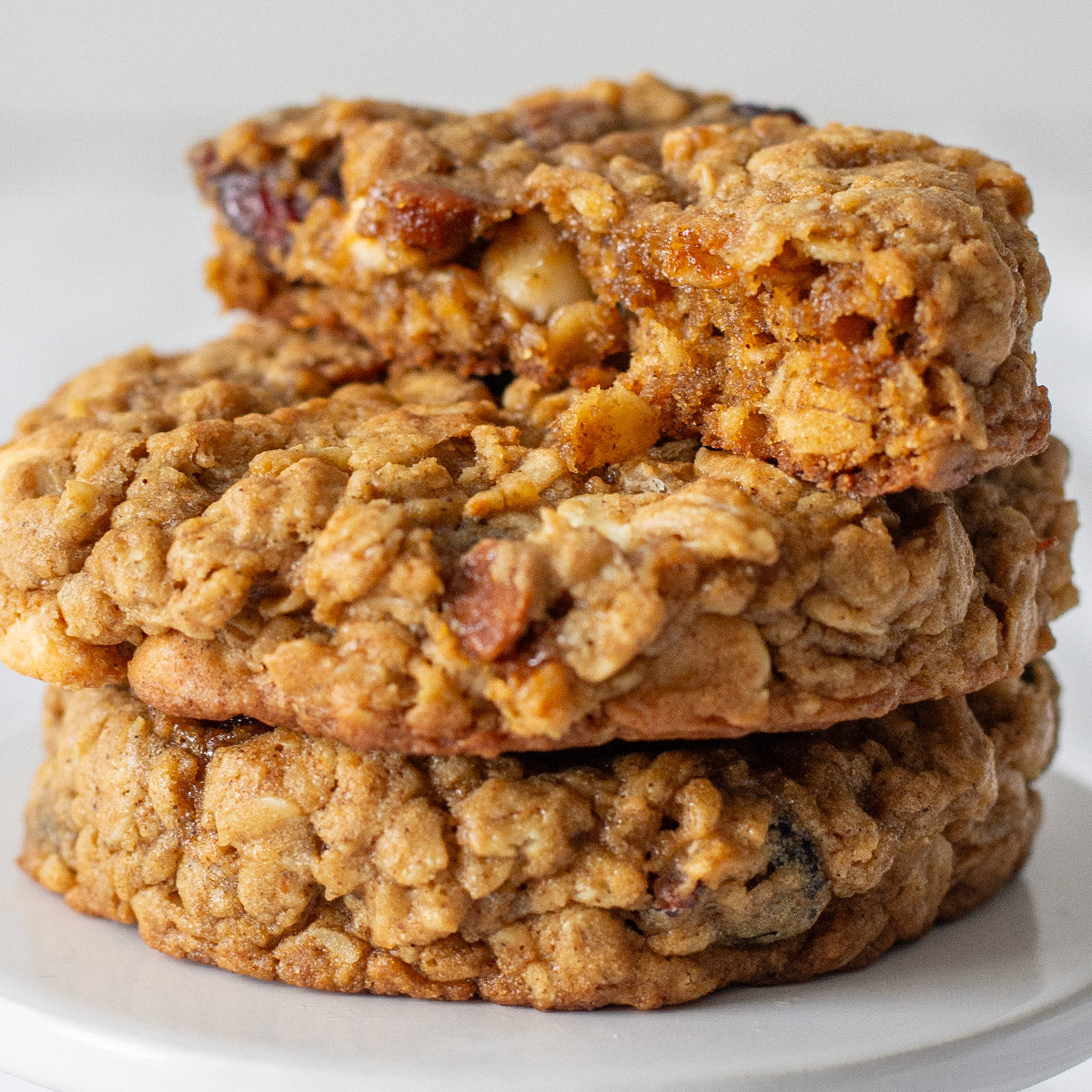 Three stacked Spiced Pumpkin Oat Cookies, with the top cookie broken in half to reveal a soft, chewy interior filled with oats, cranberries, and melted white chocolate and cinnamon chips.