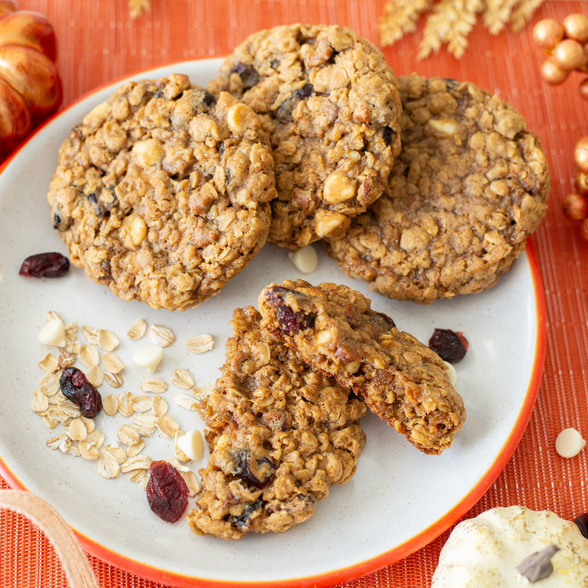 A plate of Spiced Pumpkin Oat Cookies surrounded by scattered oats, white chocolate chips, and dried cranberries, highlighting the ingredients used in the cookies.